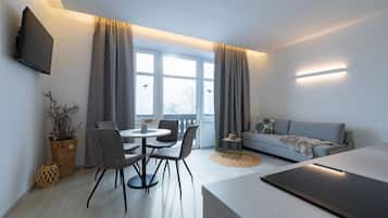 A modern living room with a dining area, a sofa, a flat-screen TV mounted on the wall, and a large window with curtains.