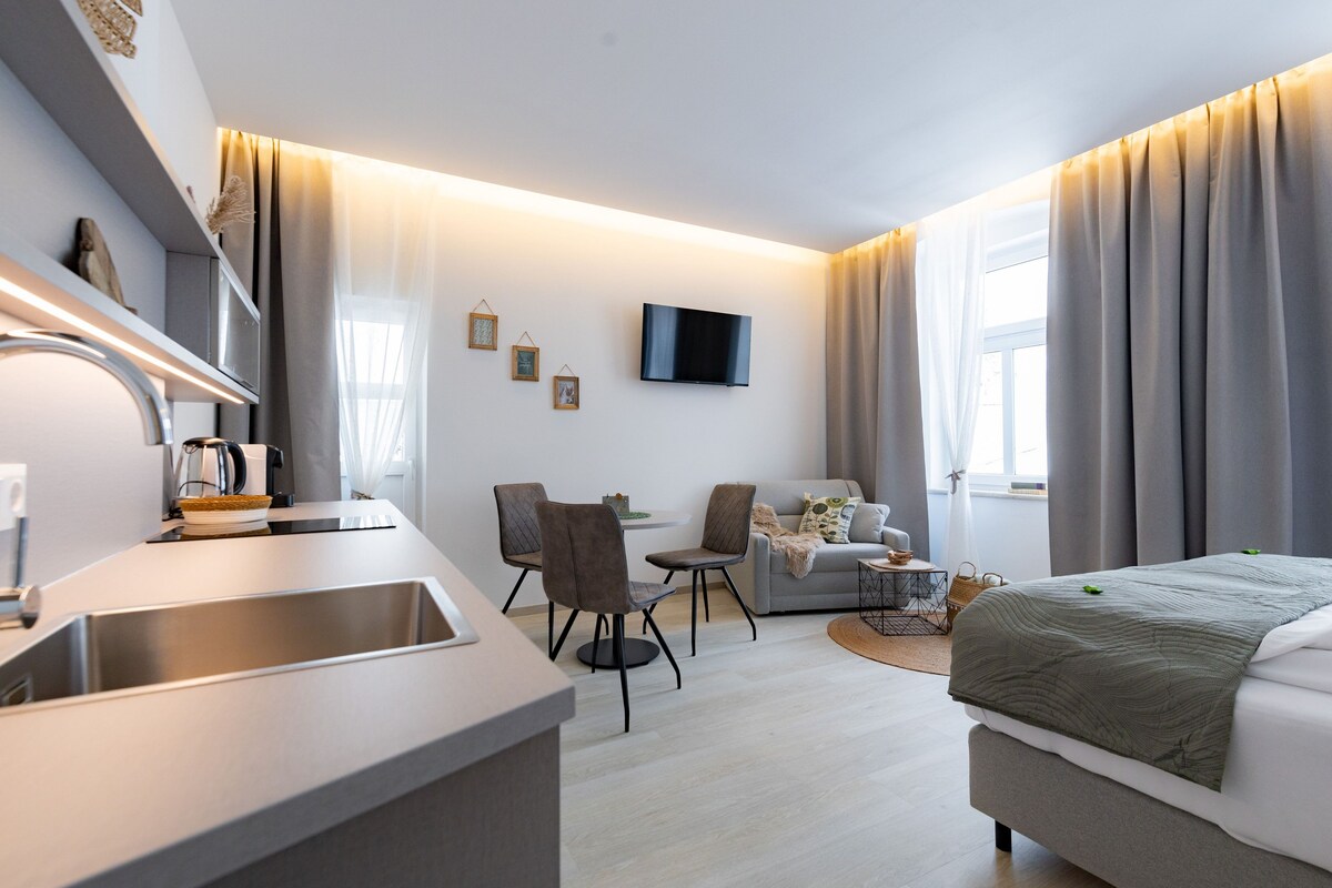 A modern hotel room with a kitchenette, a dining area, a sofa, and a bed.