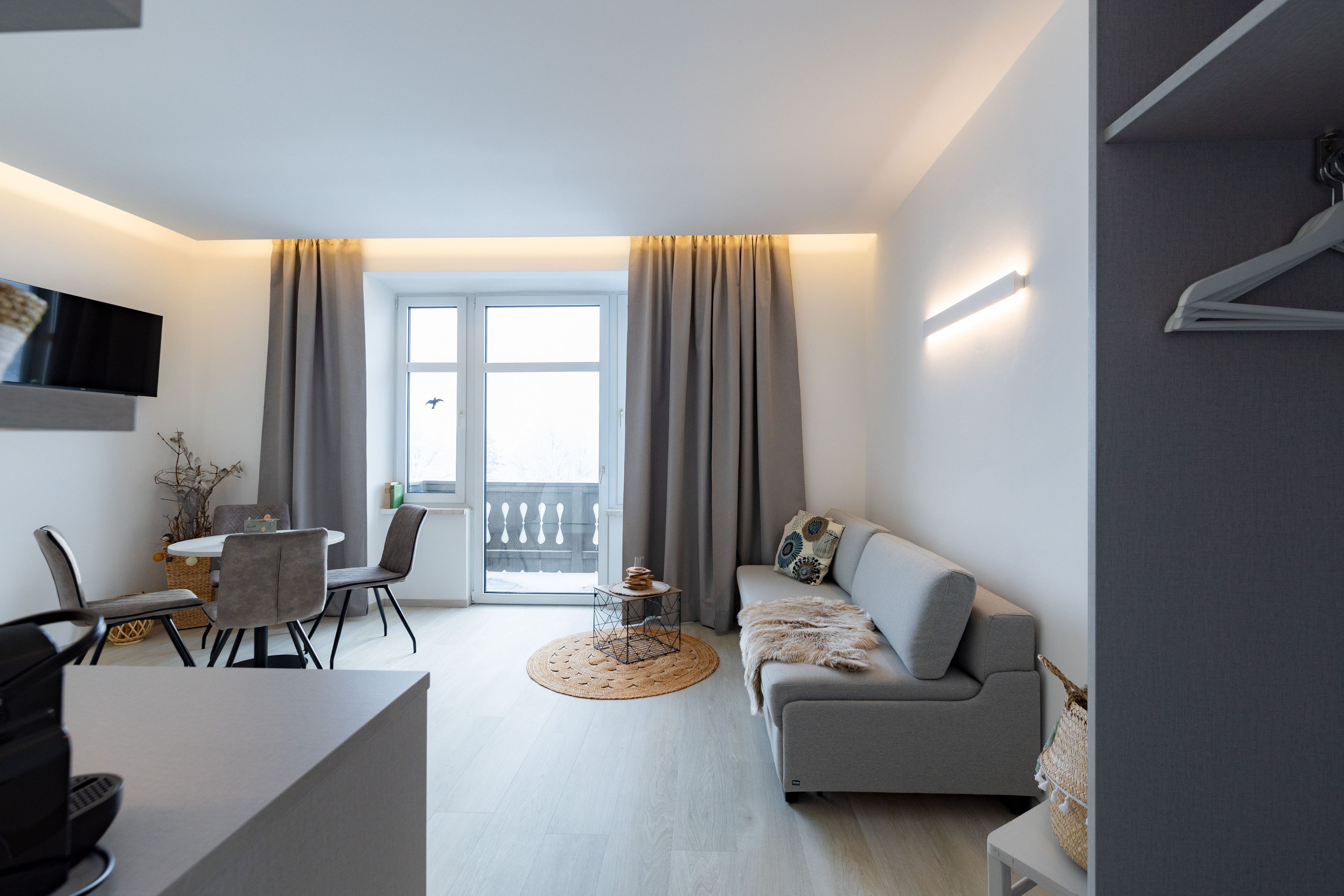 A modern living room with a grey sofa, a dining table with chairs, a flat-screen TV mounted on the wall, and a large window with curtains.