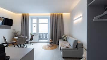 A modern living room with a grey sofa, a dining table with chairs, a flat-screen TV mounted on the wall, and a large window with curtains.