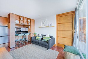 Eine moderne Küche mit Kühlschrank, Herd und Barhocker. Ein graues Sofa mit Kissen und ein Korbwannenstuhl befinden sich neben dem Fenster.