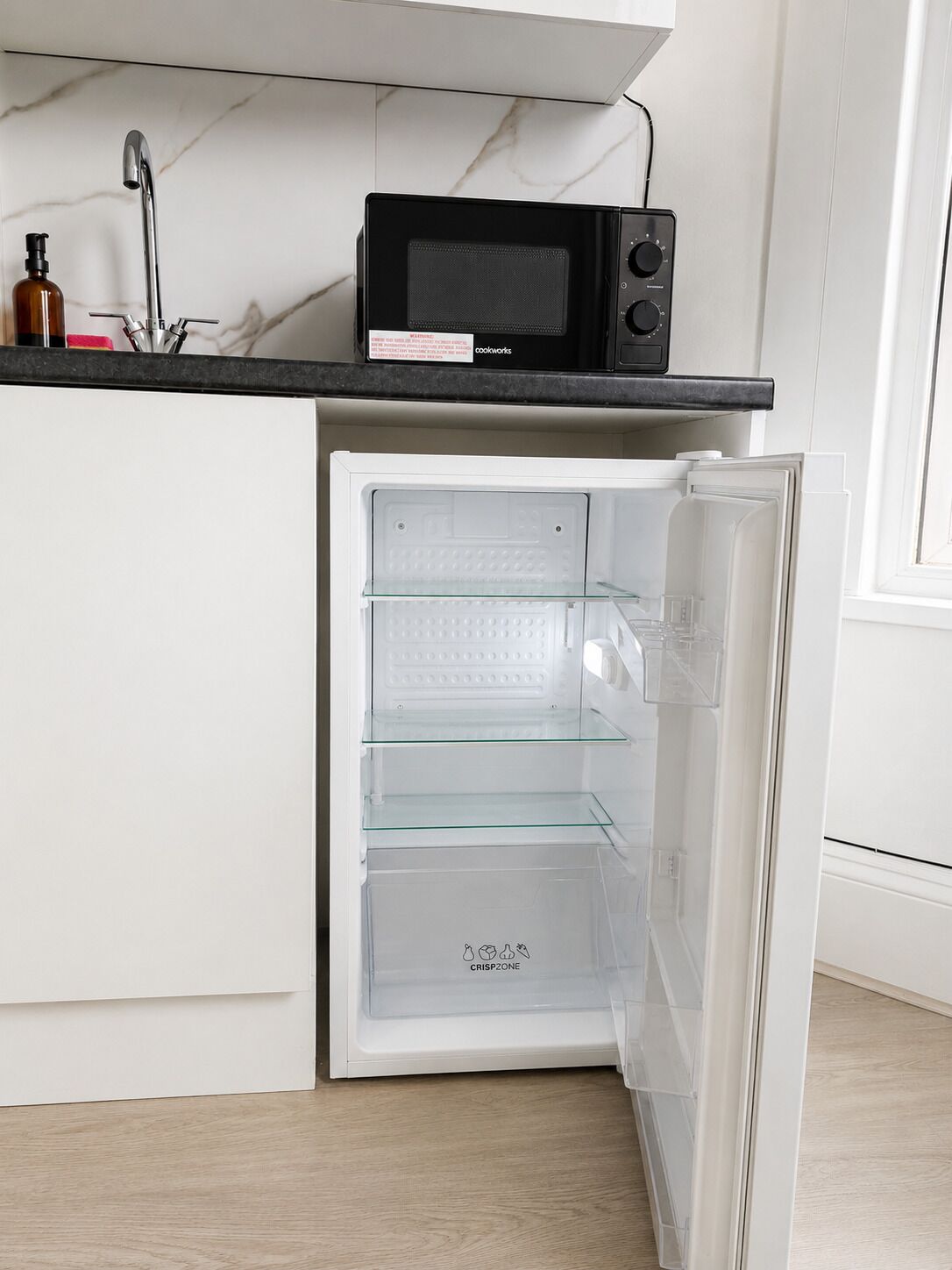 A compact kitchen with a built-in microwave, a refrigerator with a glass door, and a marble backsplash.