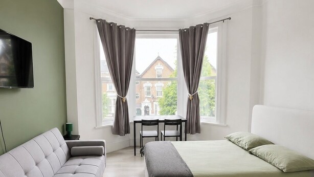 A modern bedroom with a sofa, a bed, a TV, and a view of a building through a window. - KW SELF CONTAIN STUDIOS ALEXANDRA PALACE (London)