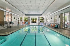 An indoor swimming pool with clear water, surrounded by lounge chairs and tables, with a tiled ceiling and glass doors leading outside. - Apartment Steps from Pentagon City Metro (Arlington)