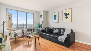 A modern living room with a black leather sofa, a wooden coffee table, a wicker chair, and a large window offering a city view. - Apartment Steps from Pentagon City Metro (Arlington)