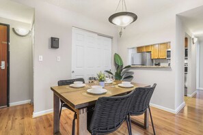 Un comedor con una mesa de madera dispuesta para cuatro, sillas negras, una planta en maceta y una cocina al fondo.