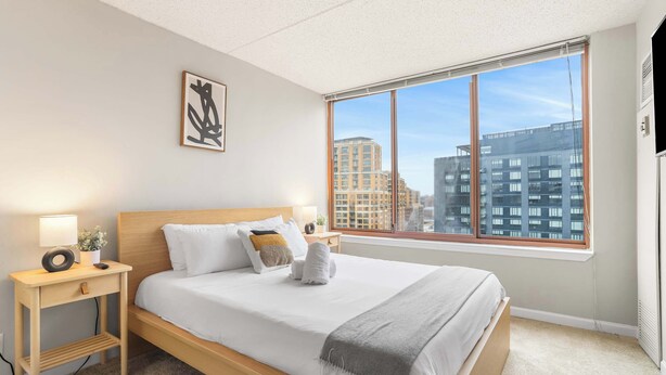 A modern bedroom with a large window, a bed with white bedding, a bedside table with a lamp, and a framed picture on the wall. - Apartment Steps from Pentagon City Metro (Arlington)