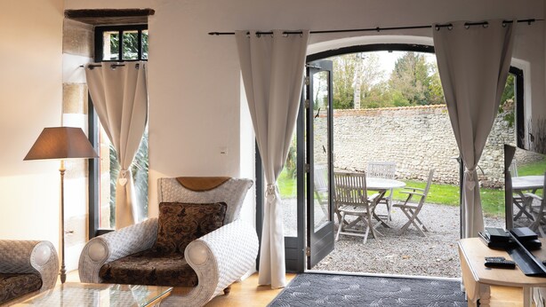A cozy living room with a sofa, armchair, and a glass coffee table. There is a door leading to an outdoor seating area with a stone wall and a bench. - Maison 2 du domaine d'embourg (Souvigny)