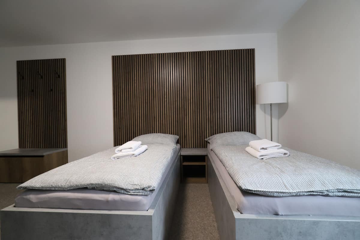 A hotel room with two single beds, a bedside table, a lamp, and a wooden panel wall.