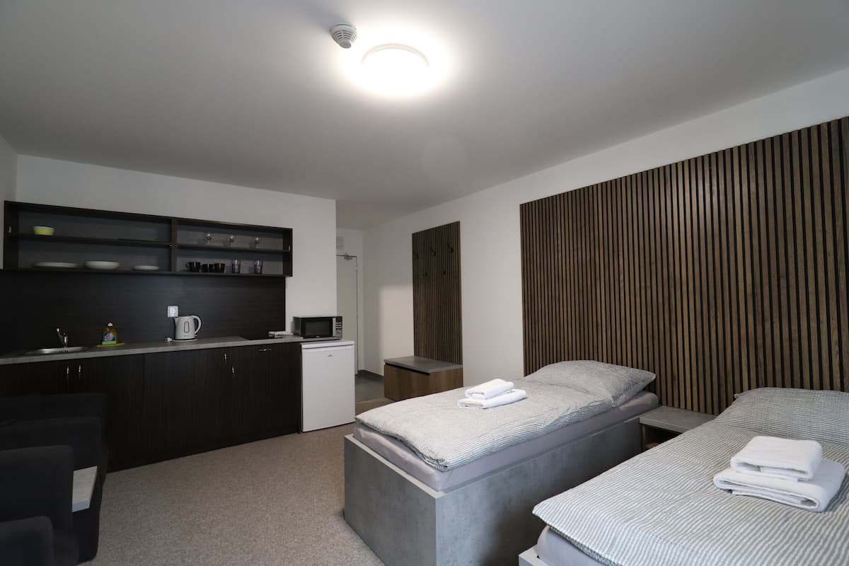 A modern hotel room with a kitchenette, two beds, a wooden panel wall, and a ceiling light.