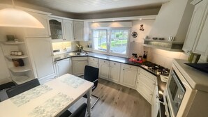 A kitchen with white cabinets, a dining table, a black chair, a stove, and a window with a view of greenery. - Apartment in Traditional Timber-Framed House with Garden in the Bergisches Land (Overath)