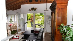 A living room with a sofa, a table, a chair, and a view of a garden through glass doors. - Apartment in Traditional Timber-Framed House with Garden in the Bergisches Land (Overath)
