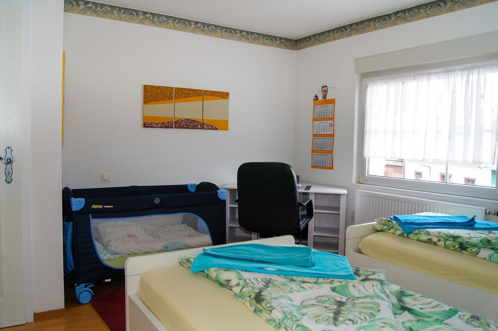 A room with two beds, a desk, a chair, a crib, and a window with curtains.