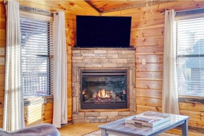 A cozy cabin interior with a stone fireplace, wooden walls, and a table with books. - Huge 3BD in TOWN! HOT TUB +Game Room +POOL Access! (Pigeon Forge)