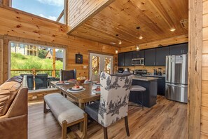 A wooden cabin interior with a dining area, kitchen, and a large window overlooking greenery. - Relaxing 5BD POOL Lodge| Theater + Hot Tub + Games (Sevierville)