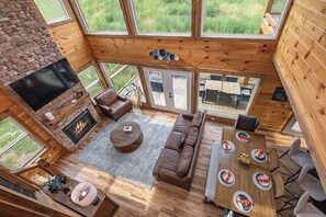 Un vaste espace de vie doté d’une cheminée, d’un canapé, d’une table à manger pour quatre personnes et de grandes fenêtres offrant une vue sur l’extérieur.