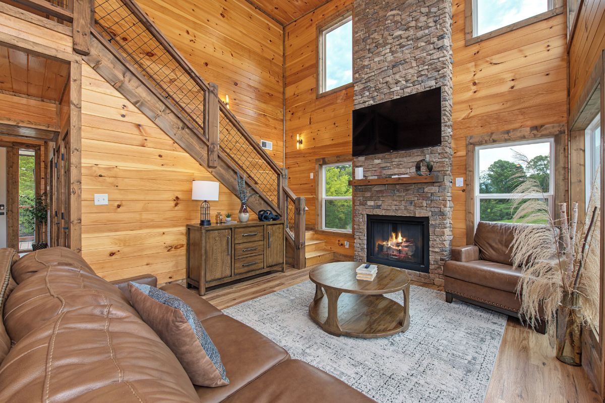 A cozy cabin interior with a stone fireplace, wooden walls, a leather sofa, and a staircase.