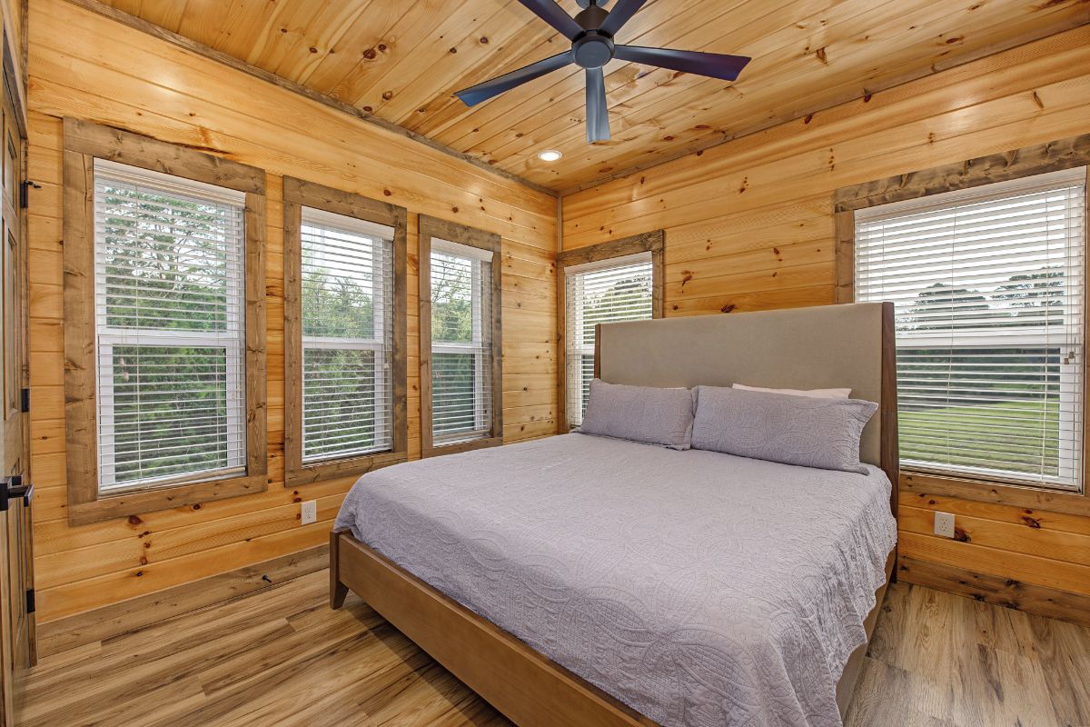 A wooden cabin bedroom with a bed, a ceiling fan, and large windows.