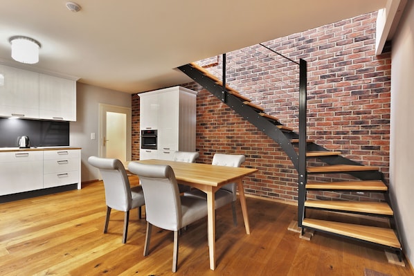A modern kitchen with white cabinets, a dining table with chairs, and a brick wall with a wooden staircase.