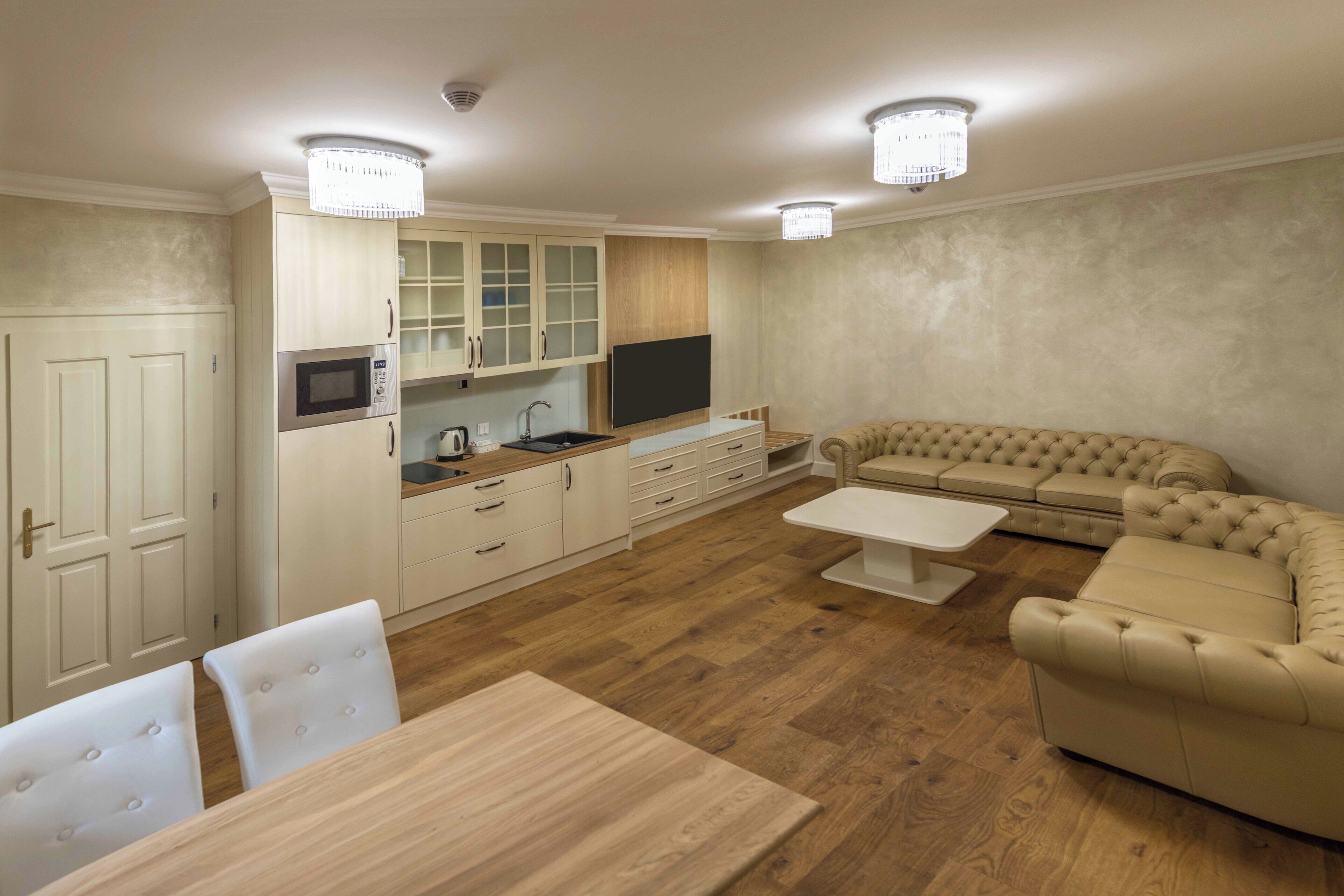 A modern kitchen with built-in appliances, a dining area with a wooden table and white chairs, and a beige sofa in a living area.