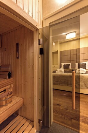 Una sauna de madera con bancos, una puerta de vidrio que da a un dormitorio con una cama y una mesita de noche.