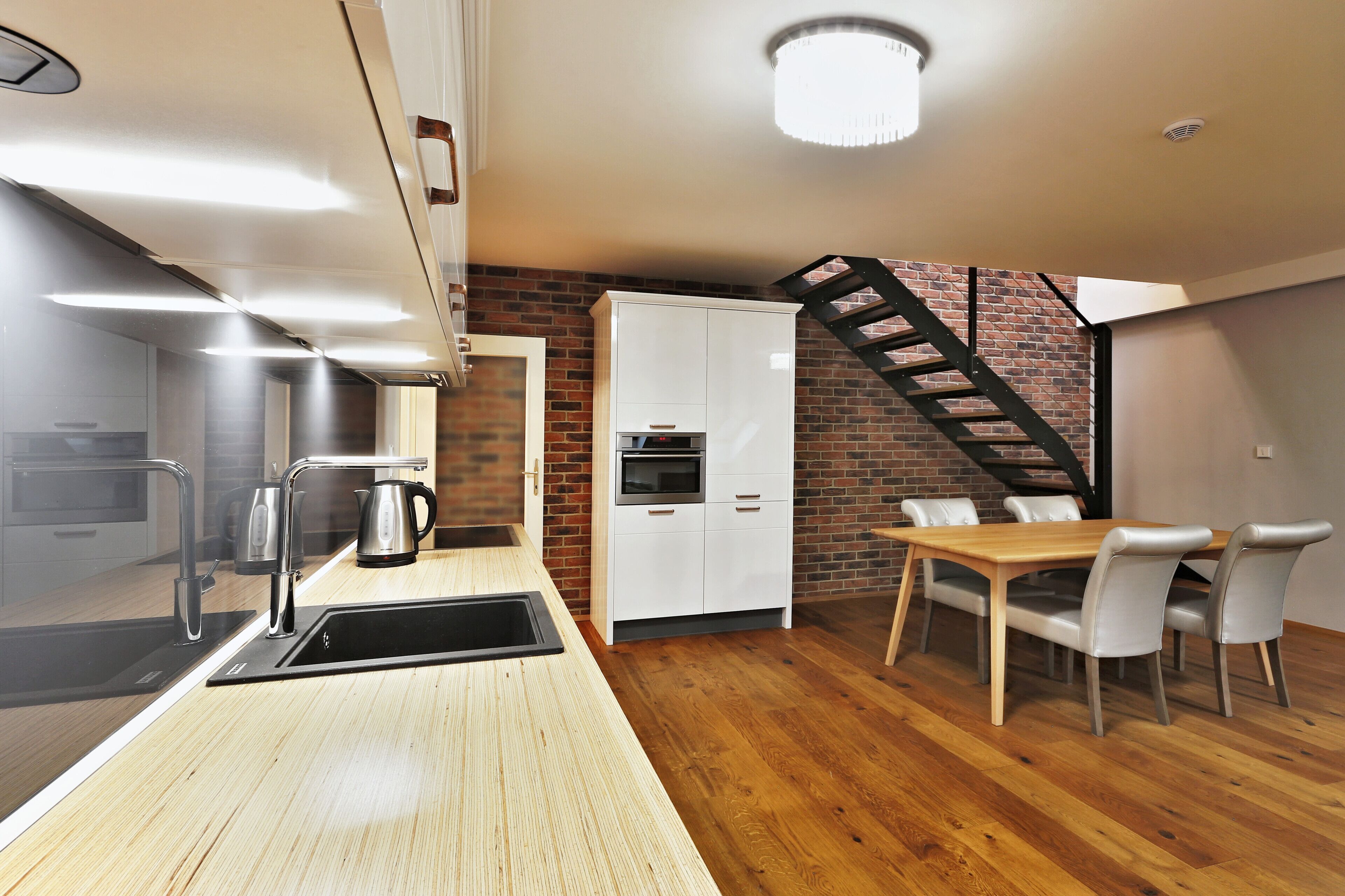 Une cuisine moderne équipée d’un évier, d’une cuisinière et de meubles blancs, un coin repas avec une table et des chaises en bois, et un mur en briques avec un escalier noir.