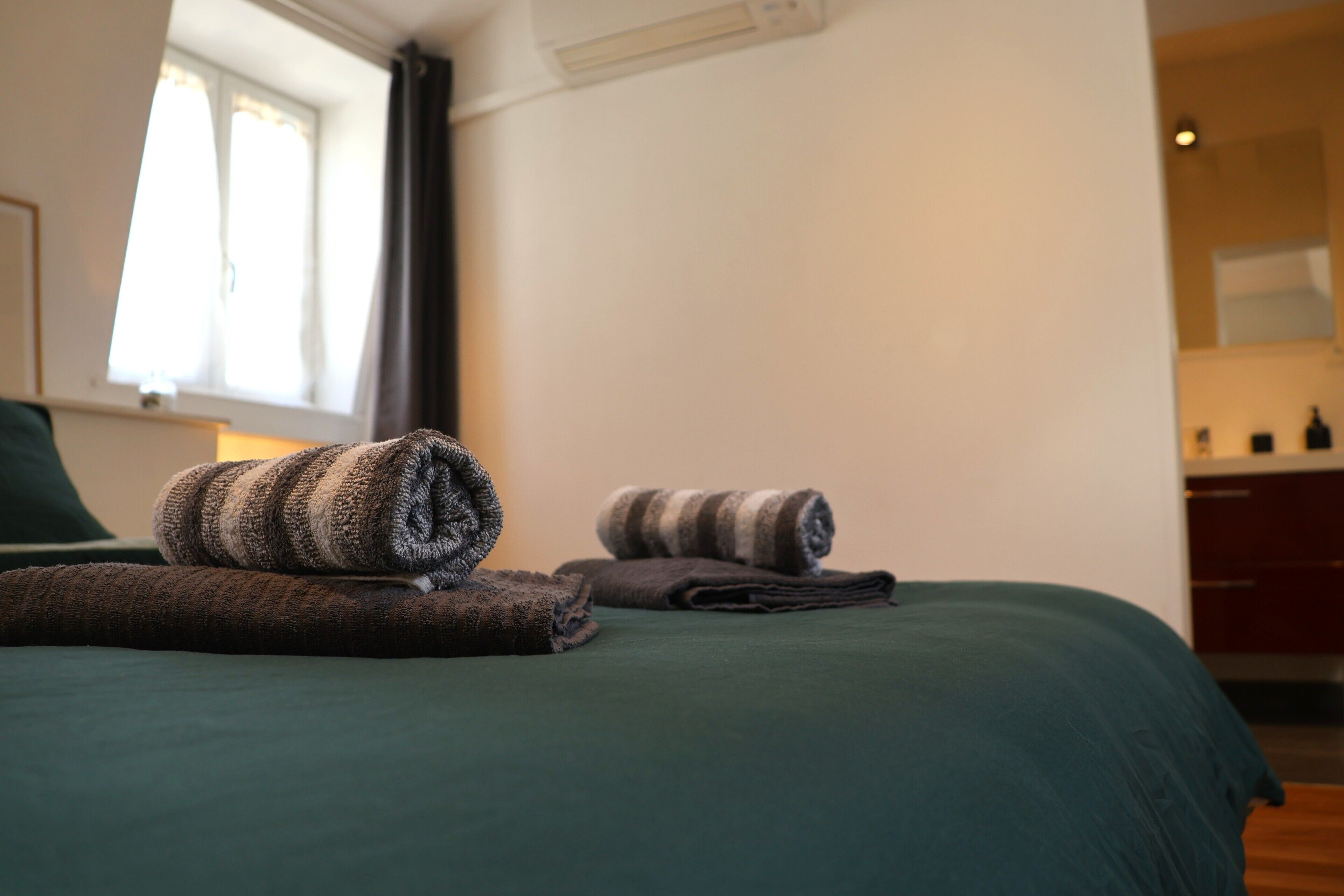 Ein ordentlich bezogenes Bett, zusammengerollte Handtücher, ein Fenster mit Vorhängen und eine Küchenzeile im Hintergrund.
