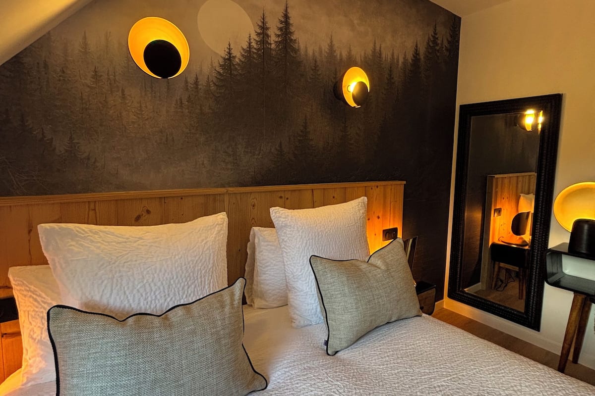 A bedroom with a sloped ceiling, a bed with white bedding, and a mural of trees and a moon.