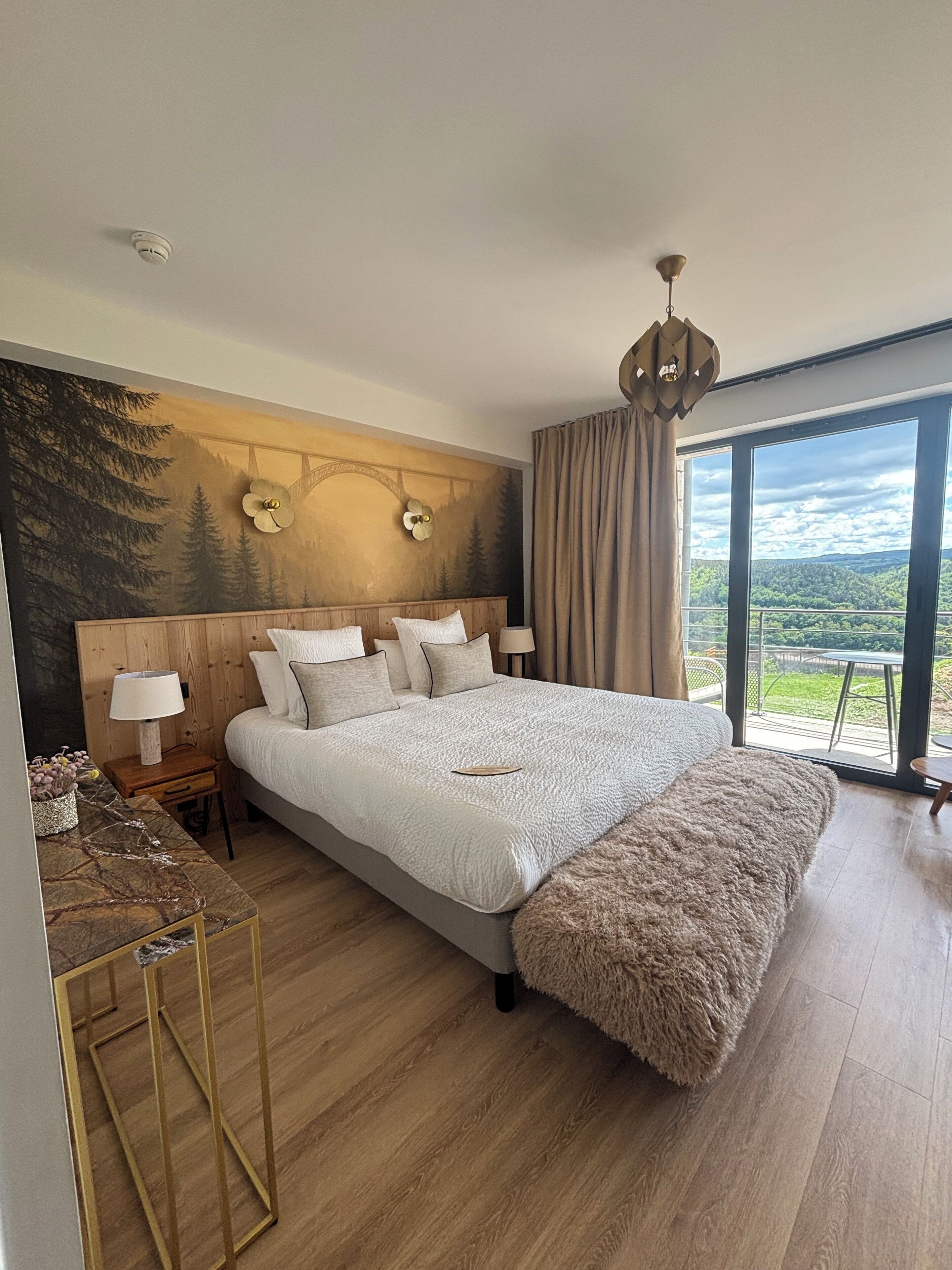 A bedroom with a large bed, a nightstand, a lamp, a painting on the wall, and a view of the outdoors.