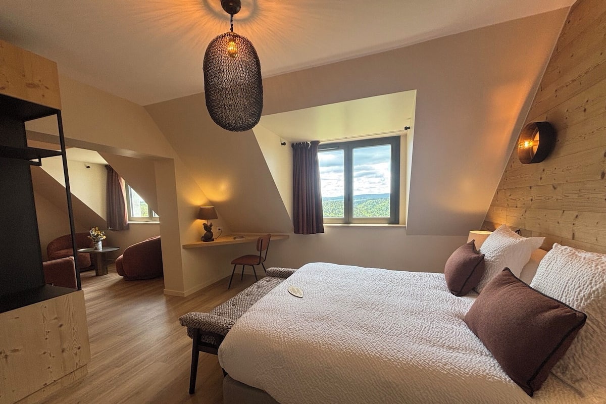 A bedroom with a large bed, a desk, and a chair, a window with curtains, and a hanging light fixture.