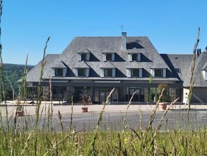 A building with a sloped roof, multiple windows, and a flat entrance area with potted plants. - Le Panoramic Garabit  (Anglards-de-Saint-Flour)