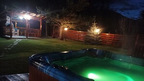 Et spabad i en baghave med en træterrasse, en stensti og en pergola med lys.