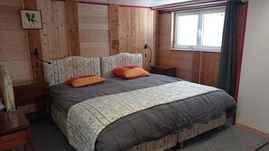 A bedroom with a wooden headboard, a bed with a patterned bedspread, two pillows, a nightstand, and a window with curtains.