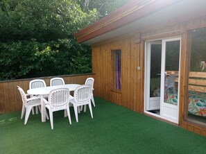 A wooden deck with a white table and chairs, a sliding glass door, and a view of a garden. - Le Cocon des 3 Pins: Mountain View, Private Terrace & Wi-Fi (Plaine)