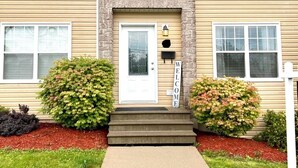 A two-story building with a beige exterior, a stone accent wall, and a white front door. There are two windows on each floor, and a small porch with steps leading to the door.