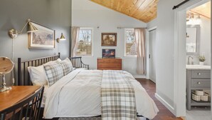 Quarto com teto de madeira, cama com colcha xadrez, cômoda, escrivaninha com abajur e janela com cortinas.