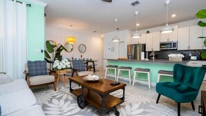 A modern living room with a sofa, a coffee table, and a teal chair. There is a kitchen area with white cabinets and a bar counter. - Storey Lake 4BR Home w/ Themed Room & Pool (Kissimmee)