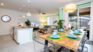 A modern kitchen with a central island, dining area with a wooden table set for six, and a large window providing natural light. - Storey Lake 4BR Home w/ Themed Room & Pool (Kissimmee)