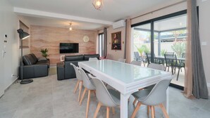 A modern living room with a glass dining table, grey chairs, a sofa, a TV, and a large window with curtains. - Prestigious gîte for 24 people - ideal for companies or large groups (Tignieu-Jameyzieu)