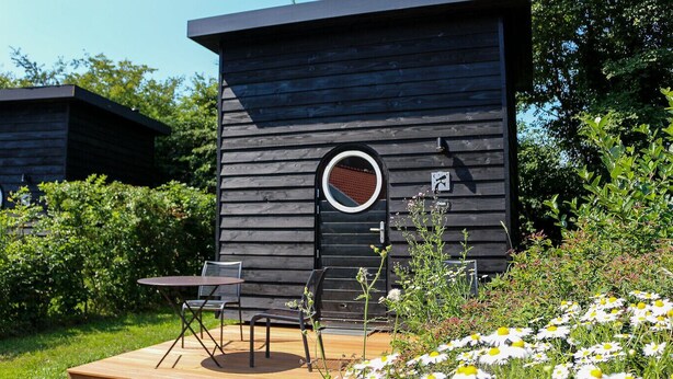 Una cabana de fusta moderna amb una finestra circular, una terrassa amb mobles d'exterior i un jardí ple de margaridades.