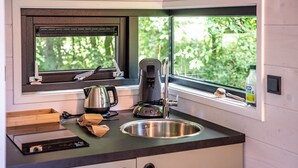 Een kleine keuken met een roestvrijstalen spoelbak, een waterkoker, een koffiezetapparaat en een raam met uitzicht op groen.