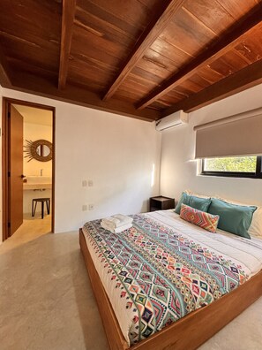 Ein Schlafzimmer mit Holzboden, einem Bett mit bunter Decke, einem Nachttisch und einem Fenster mit Blick auf Grünflächen.