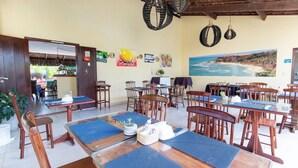 Ein Restaurant mit Holztischen und -stühlen, blauen Platzsets, hängenden Lampen und einem Wandbild mit einer Strandansicht.