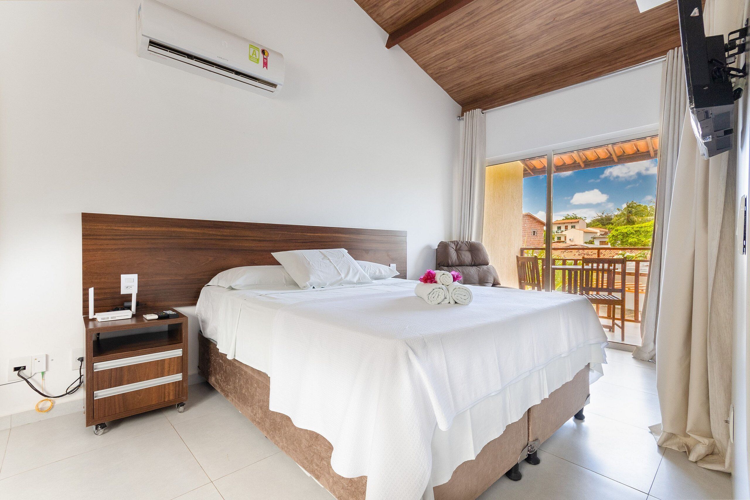 Camera da letto con una testiera in legno, copripiumino bianco, comodino con lampada e vista all'esterno da una grande finestra.