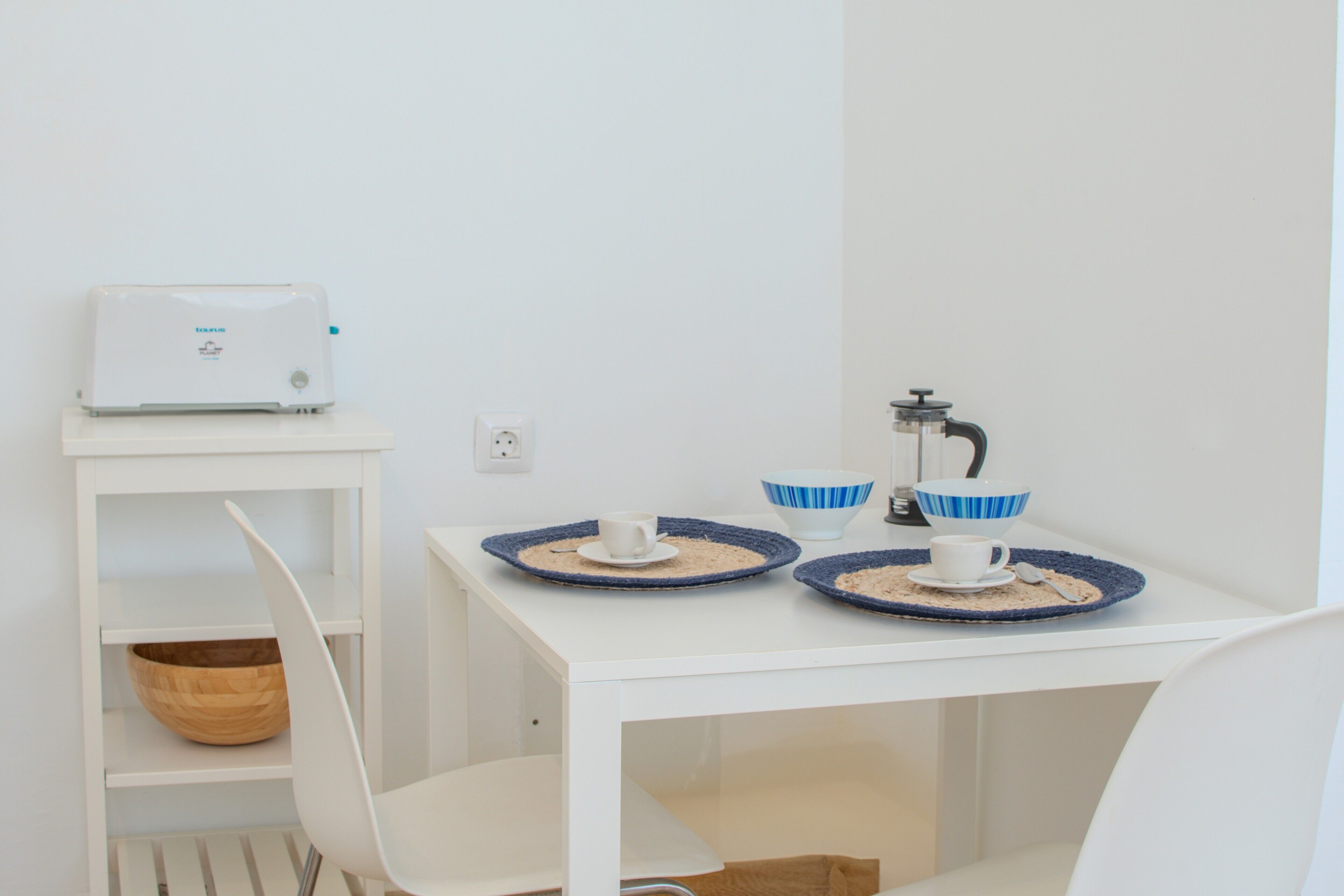 Une table blanche avec une machine à café, deux tasses à bordure bleue et un bol en bois, le tout posé sur une étagère blanche.