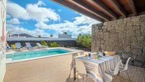 A poolside area with a table and chairs, a stone wall, and a view of the sky and mountains.