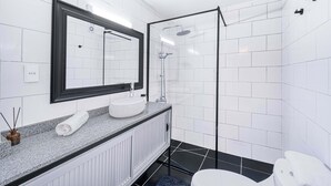 Un baño moderno con pared de azulejos blancos, un espejo grande, un lavamanos blanco y una ducha con mampara de cristal.