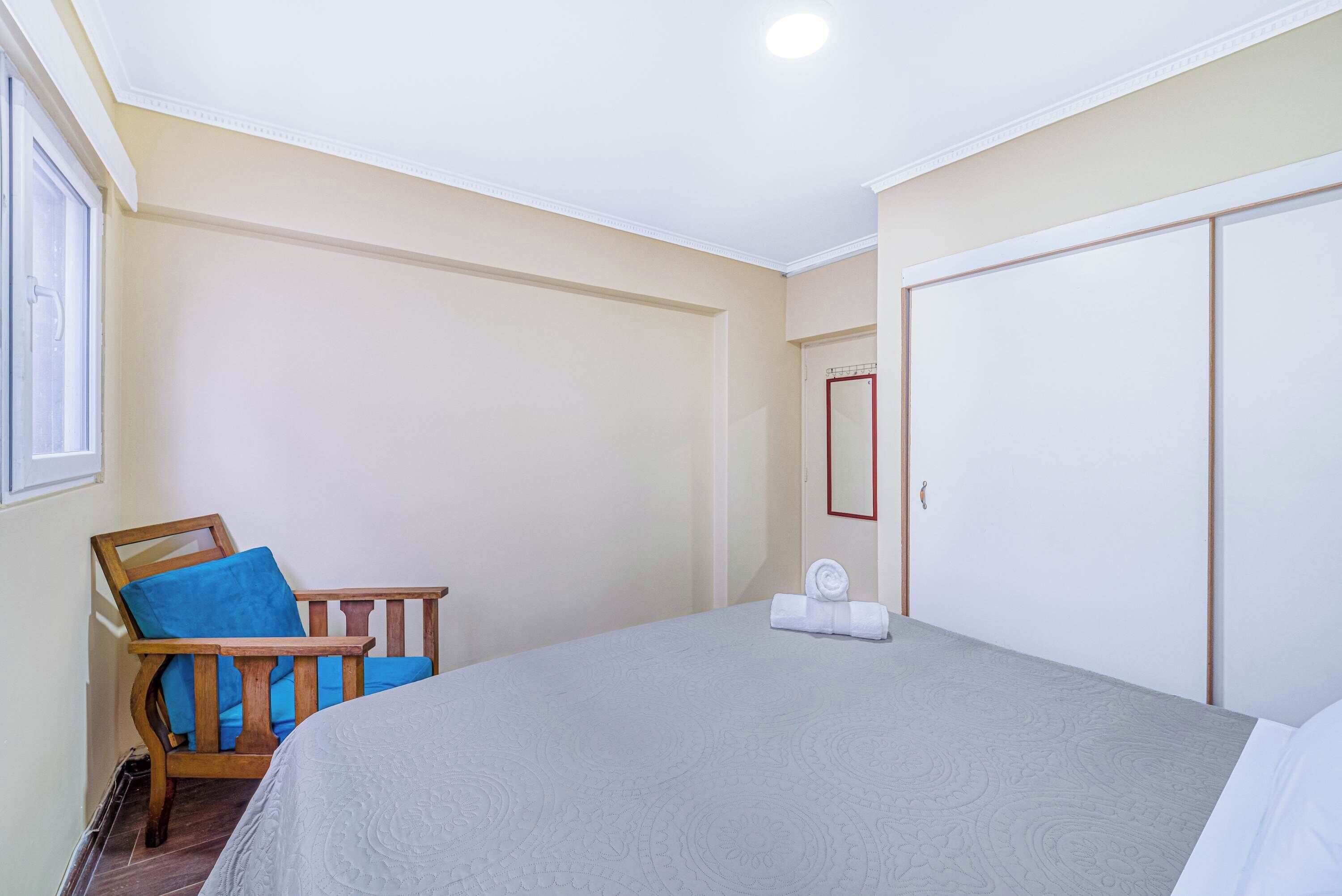 Ein Schlafzimmer mit einem Bett, einem Stuhl mit blauer Sitzkissen, einem Kleiderschrank und einem Fenster.