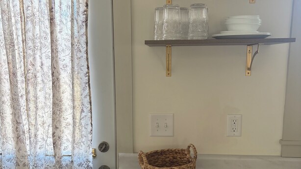 Et køkken med en marmor bordplade, en fletkurv, en vægmonteret hylde med glas og en dør med blomstret gardin.