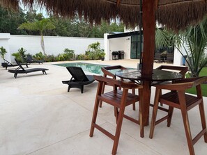 An outdoor pool area with lounge chairs, a wooden table, and a thatched roof structure.